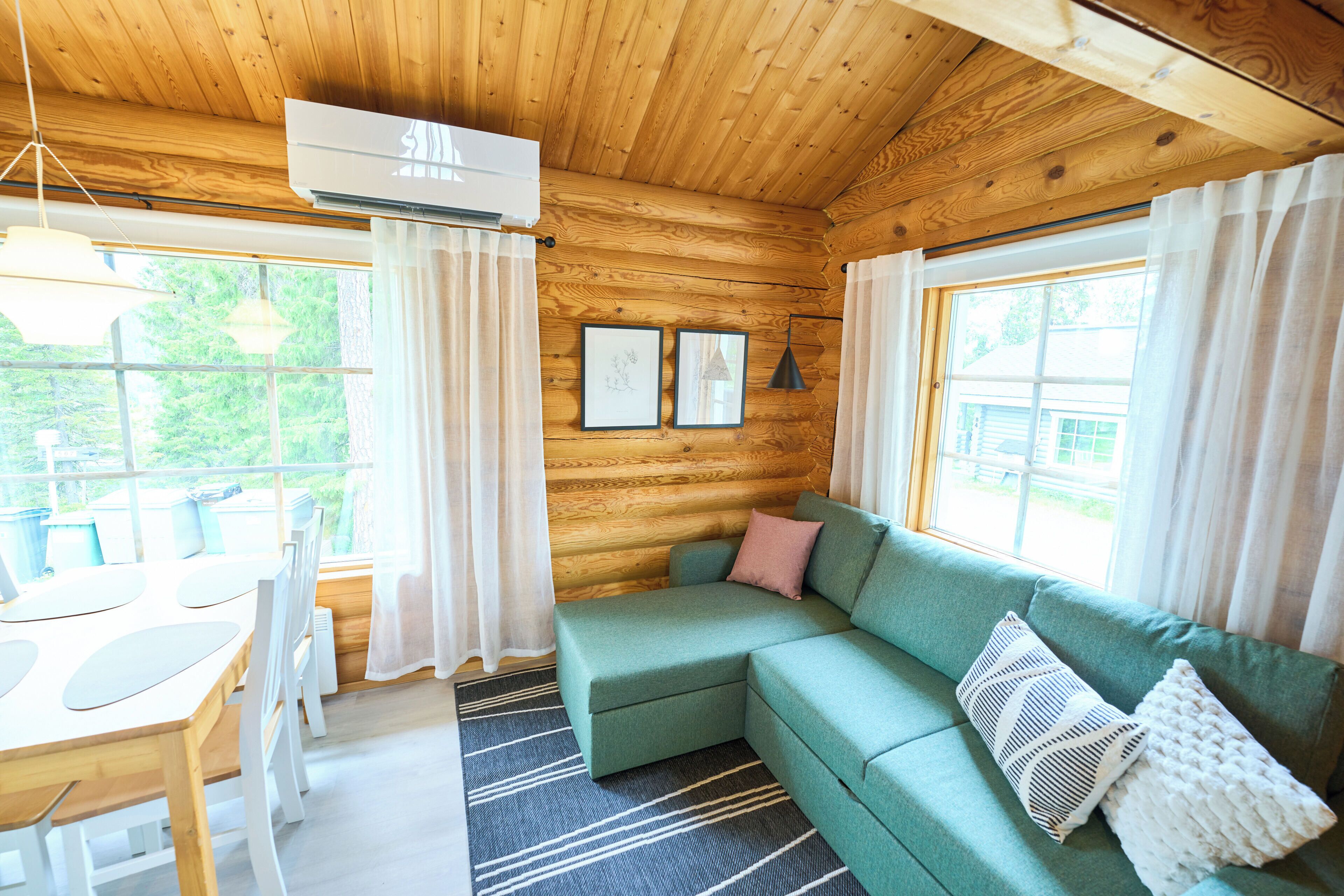 A wooden cabin interior with a green sofa, a dining table, and a large window.