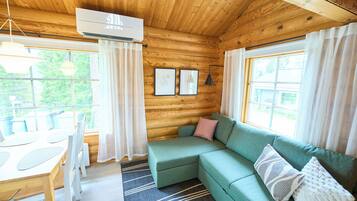 A wooden cabin interior with a green sofa, a dining table, and a large window.