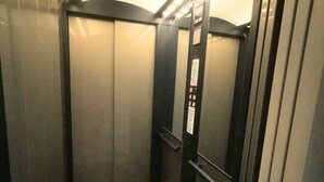 Interior de elevador com paredes metálicas, painel de comando com botões e teto refletivo.