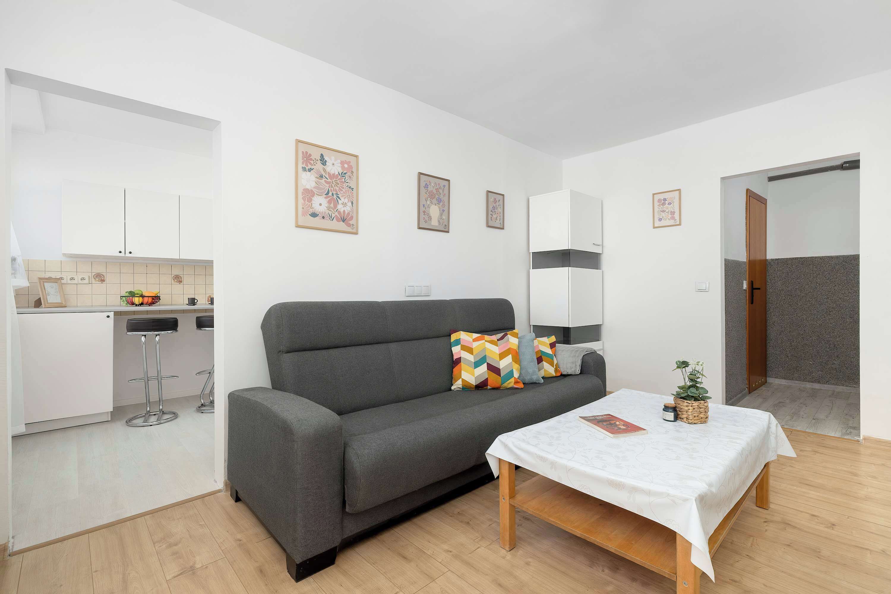 En moderne stue med en grå sofa, et lille bord med hvid dug, og et køkkenområde med hvide skabe samt en skål med frugt.