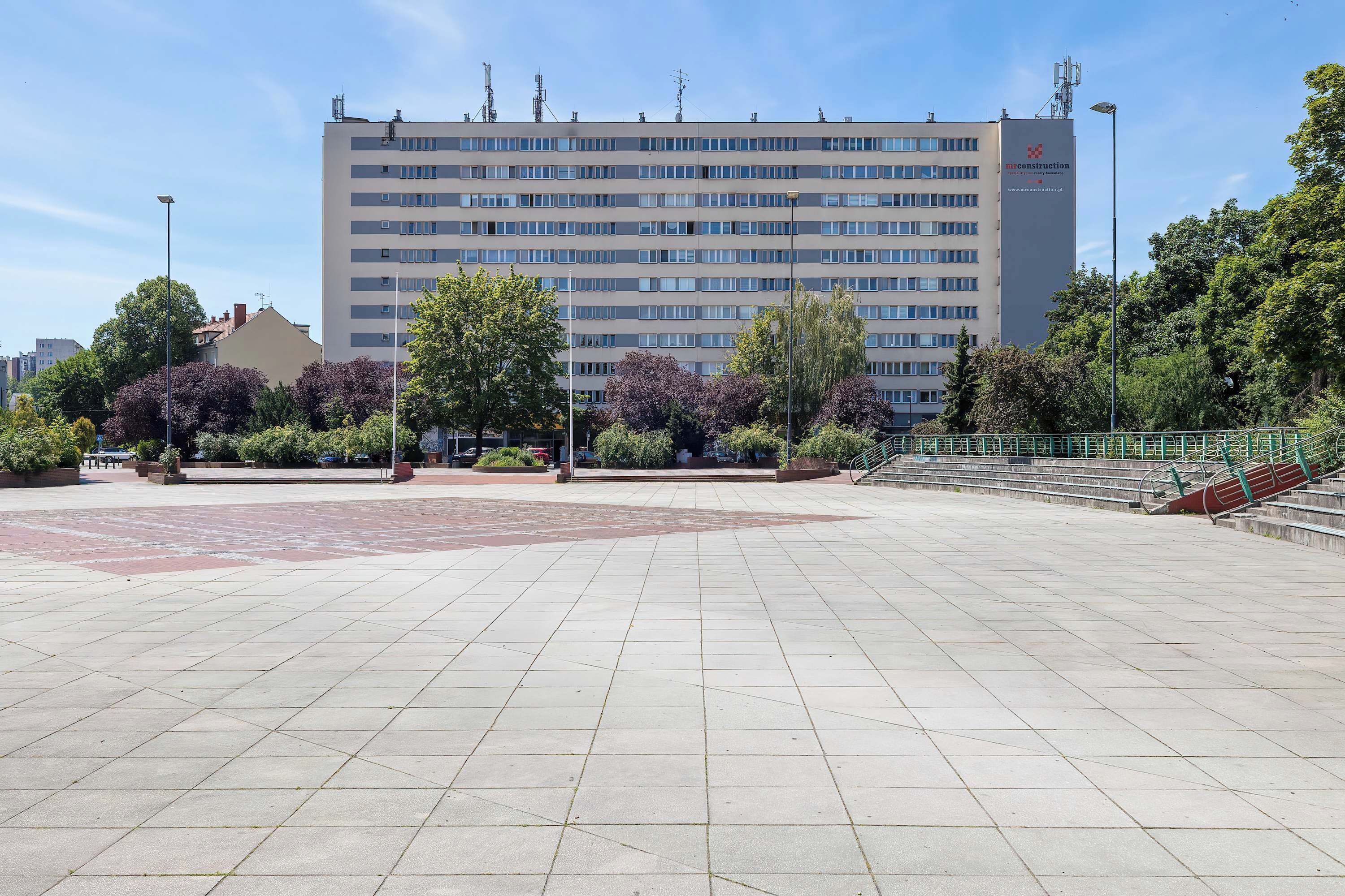 A large, multi-story building with a flat facade, surrounded by trees and a paved plaza.