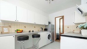 A modern kitchen with white cabinets, a washing machine, two bar stools, and a countertop with a fruit bowl and kettle. - Cozy Apartment for 3 People | Great Location (Gliwice)