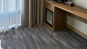 A hotel room with a large window, a flat-screen TV, a wooden desk, and a small refrigerator.