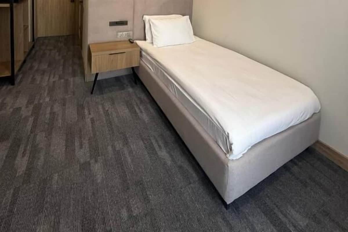Una cama individual en una habitación con alfombra gris, un armario de madera y una pared de acento naranja.