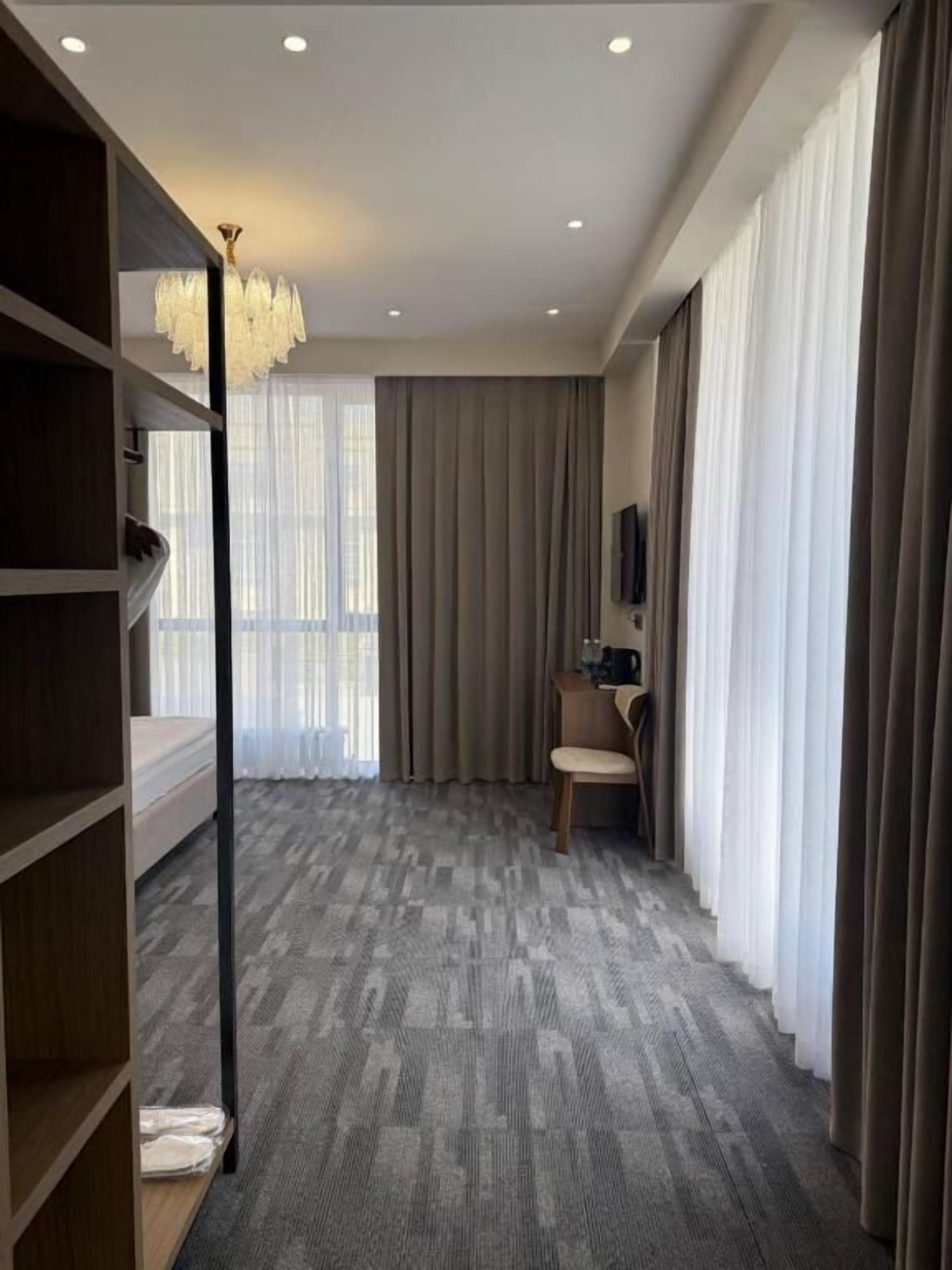 A hotel room with a carpeted floor, a bookshelf, a desk with a chair, a television, and a large window with curtains.