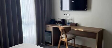 A hotel room with a bed, a desk with a chair, a flat-screen TV, and a window with curtains.