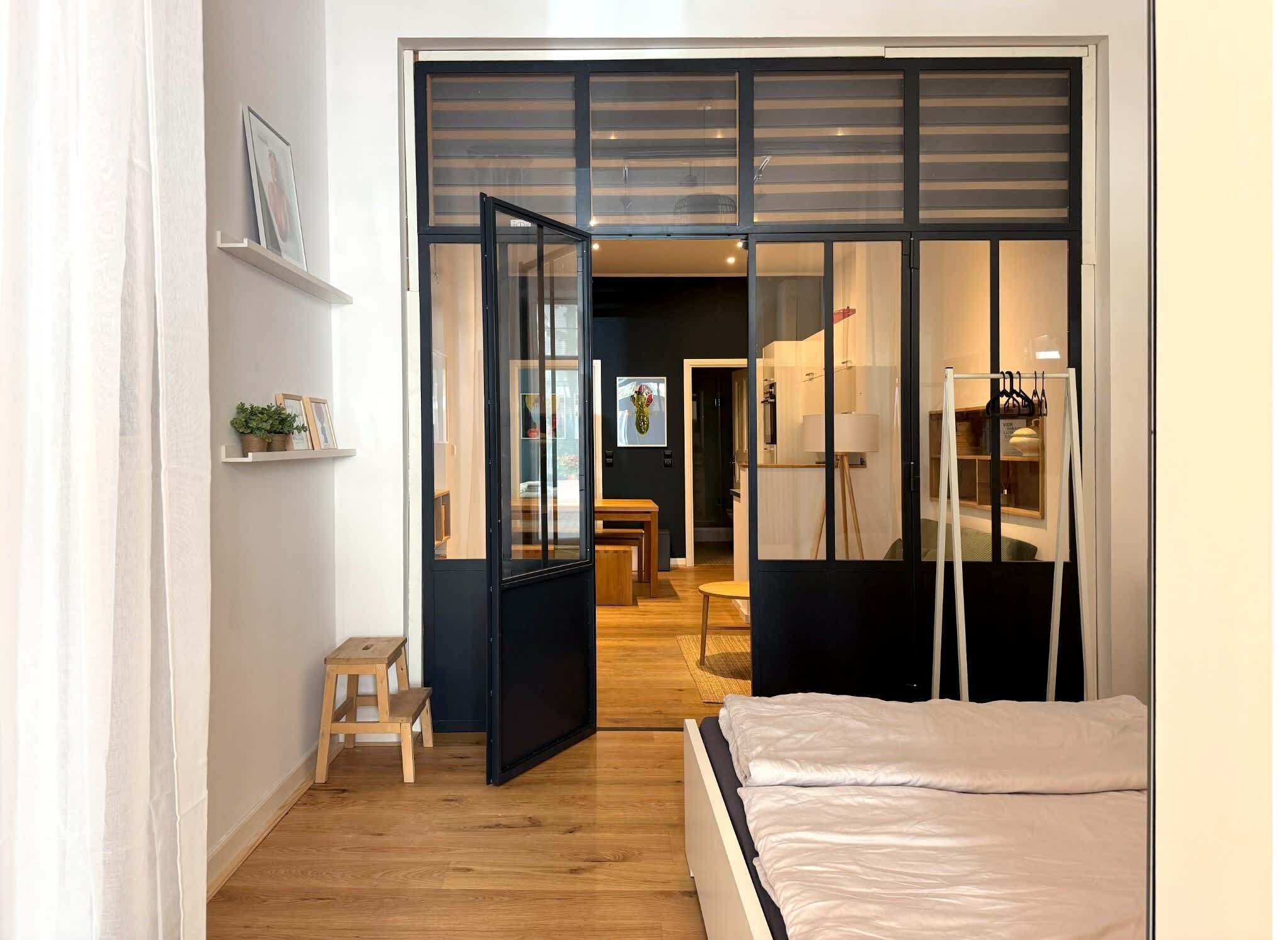 Ein Schlafzimmer mit einem Bett, einem kleinen Hocker, einem Regal mit Pflanzen und einer Schiebetür zu einem Essbereich.