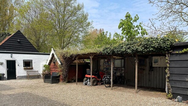Een grindpad dat leidt naar een wit gebouw met een zwart dak, een overdekt buitengebied met een fiets, en een rood voertuig dat er geparkeerd staat.