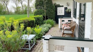 Een terras in de tuin met een tafel en stoelen, een wit gebouw met een zwarte deur, en een haag op de achtergrond.