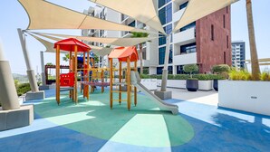 Ein Spielplatz mit Klettergerüsten und Rutschen, umgeben von modernen Gebäuden und Palmen.