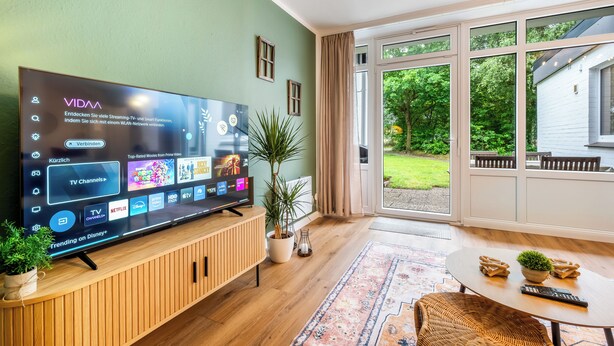 Una sala de estar moderna con un televisor de pantalla plana grande, muebles de madera, una planta en maceta y vistas al exterior.