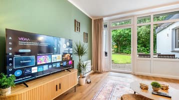 Ein modernes Wohnzimmer mit großem Flachbildfernseher, Holzmöbeln, einer Zimmerpflanze und Blick ins Freie.