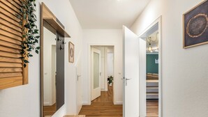 Un pasillo con piso de madera, un espejo, una planta y una puerta que da a una habitación con pared verde y un sofá.