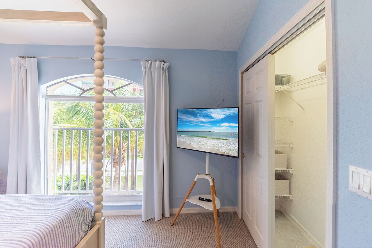 A bedroom with a bed, a TV on a stand, a window with curtains, and a view of palm trees.