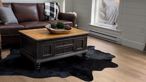Una sala de estar con un sofá de cuero marrón, una mesa de centro de madera con adornos, un tapete de pelo alto negro y un televisor montado en la pared.