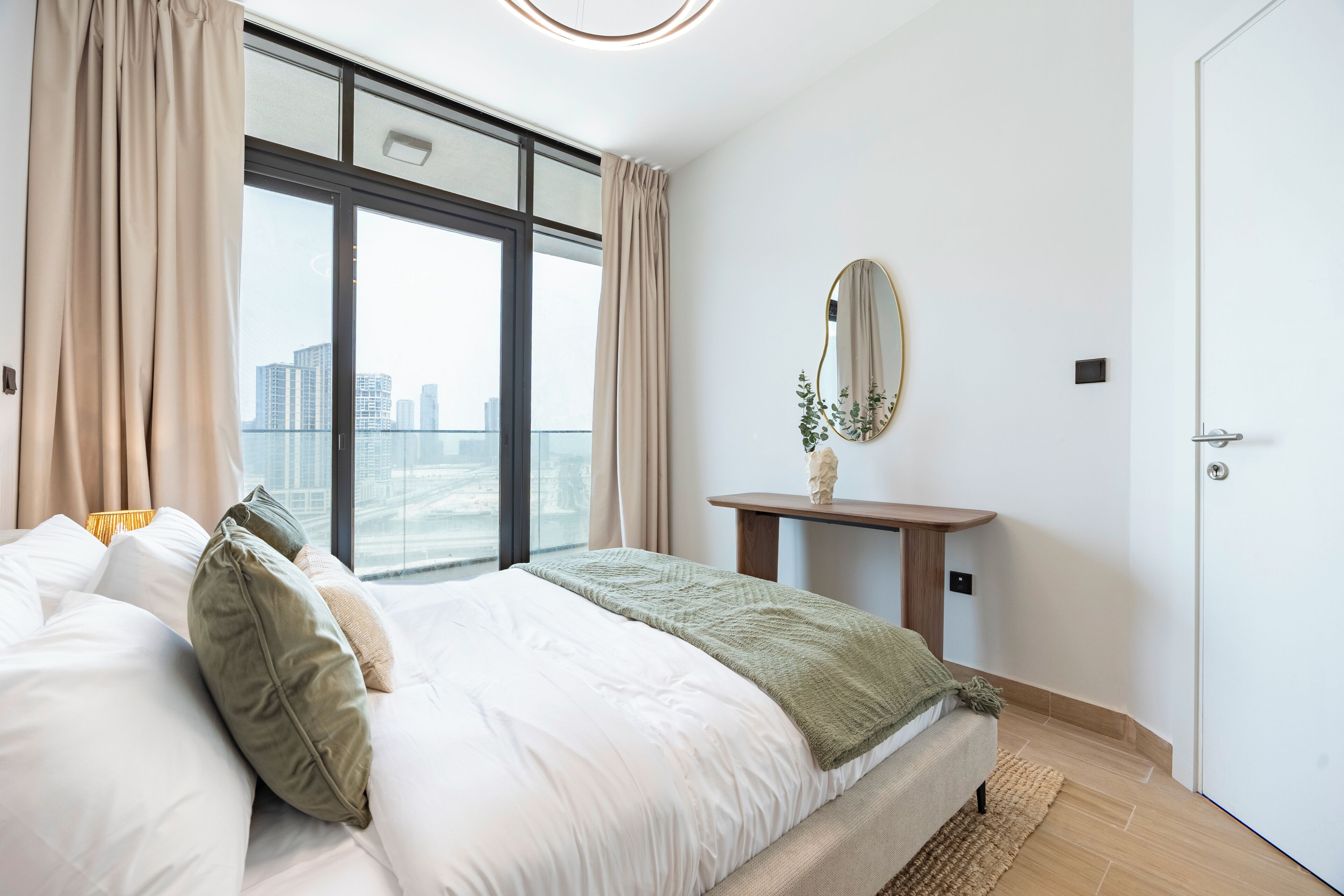 A bedroom with a bed, a bedside table with a mirror, a view of the city through large windows, and a door.