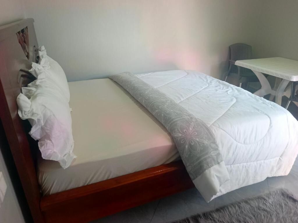 Uma cama de solteiro com estrutura de madeira, colchão branco e edredom branco com padrão floral. Um pequeno criado-mudo com superfície branca está à direita da cama.