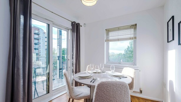 Un coin repas avec une table dressée pour quatre personnes, une fenêtre avec des stores et une vue sur un bâtiment à l’extérieur.