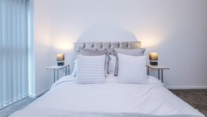 Une chambre d’hôtel avec un lit, deux tables de chevet avec des lampes, une tête de lit et une fenêtre avec des stores.