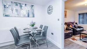 A modern living room with a glass dining table, grey chairs, a black clock, and a large abstract painting. - 3BR Elegant House in Leeds | Sleeps 6 | Driveway (West Yorkshire)