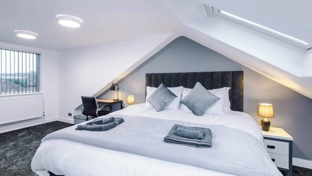 A modern hotel room with a sloped ceiling, a large bed with pillows, a desk, and a chair. - 3BR Elegant House in Leeds | Sleeps 6 | Driveway (West Yorkshire)