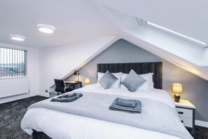 Quarto de hotel moderno com teto inclinado, uma cama grande com travesseiros, uma escrivaninha e uma cadeira.