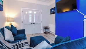 Ett rum med en blå accentvägg, en platt-TV som är monterad på väggen, en vit dörr med persienner, en lampa på ett sidobord och en soffa med kuddar.