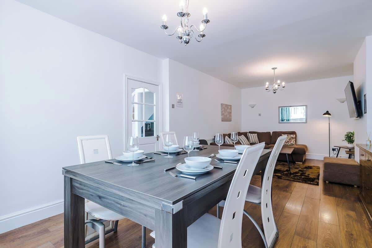 A dining area with a wooden table set for four, white chairs, a chandelier, a sofa, and a TV mounted on the wall.