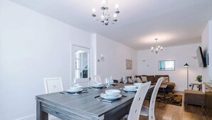 En matplats med ett träbord för fyra personer, vita stolar, en kristallkrona, en soffa och en tv som är monterad på väggen.
