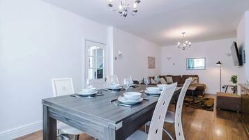 A dining area with a wooden table set for four, white chairs, a chandelier, a sofa, and a TV mounted on the wall.