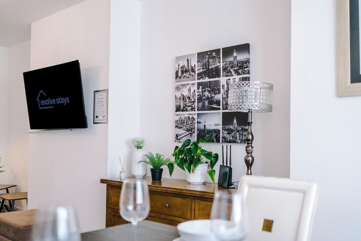 A modern living room with a flat-screen TV, a cityscape wall art, a wooden cabinet with plants, and a dining area with a table and chairs.