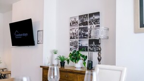Ett modernt vardagsrum med en platt-TV, en väggdekoration som föreställer en stadsbild, ett träskåp med växter och en matplats med ett bord och stolar.