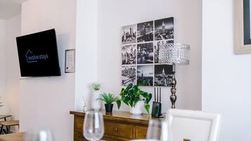 A modern living room with a flat-screen TV, a cityscape wall art, a wooden cabinet with plants, and a dining area with a table and chairs.
