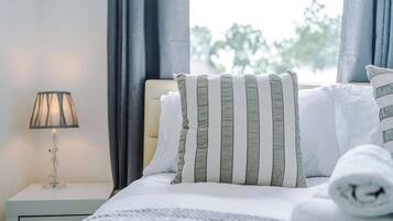 A neatly made bed with striped pillows, a white blanket, and a rolled towel on a nightstand with a lamp.