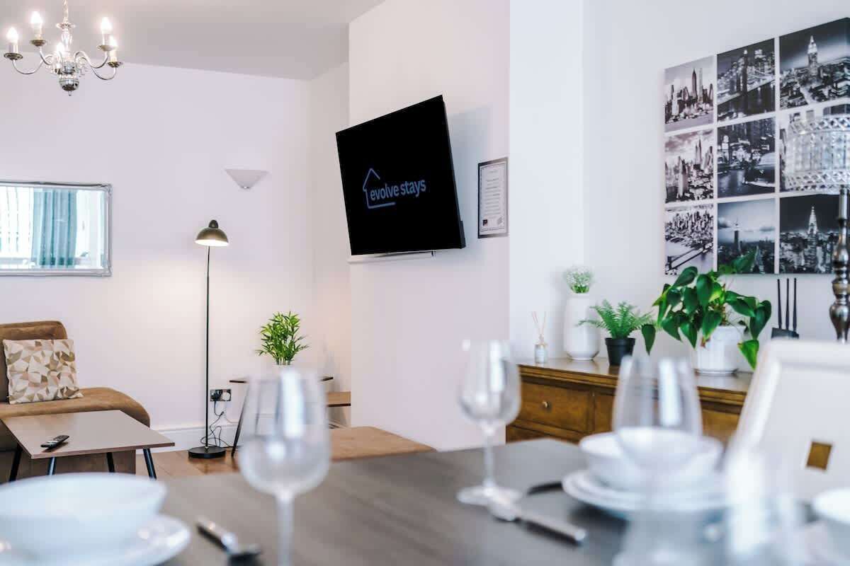 Une chambre d’hôtel moderne équipée d’un téléviseur à écran plat, d’un coin repas avec une table dressée pour quatre personnes, d’un canapé et d’un lustre.