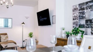 Ett modernt hotellrum med en platt-TV, en matplats med ett bord för fyra personer, en soffa och en kristallkrona.