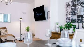 A modern hotel room with a flat-screen TV, a dining area with a table set for four, a sofa, and a chandelier.