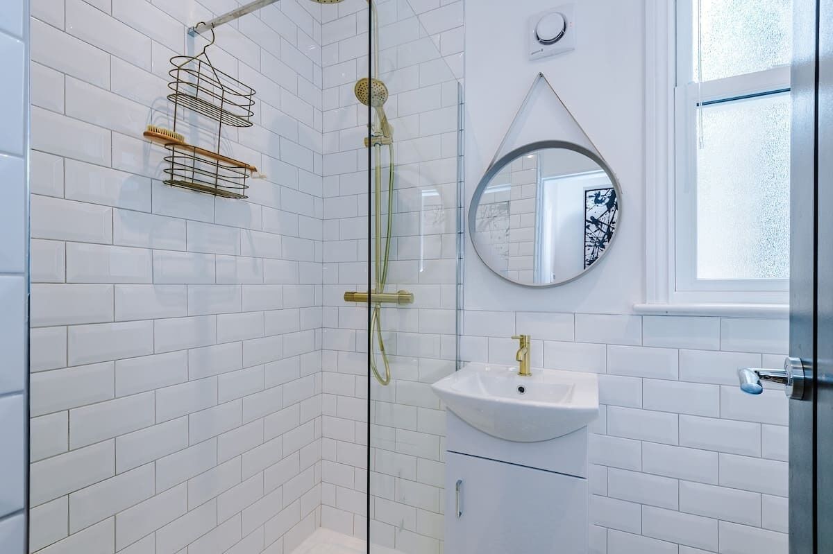 A modern bathroom with white tiled walls, a glass-enclosed shower, a white sink, and a round mirror.