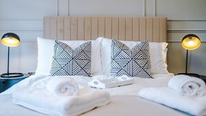 Een netjes opgemaakt bed met geometrische kussens, een nachtkastje met een lamp, en een dienblad met een glas.