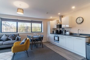 A modern kitchen with white cabinets, a dining table with chairs, a sofa, and a large window with a view of a fence. - Spacious Salford Quays Apartment, Prime Location (Greater Manchester)