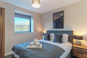 A bedroom with a bed, a window, a nightstand, a lamp, and a painting on the wall. - Spacious Salford Quays Apartment, Prime Location (Greater Manchester)