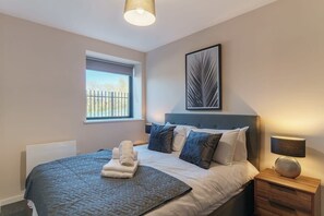 A bedroom with a bed, a bedside table, a lamp, a window, and a framed artwork on the wall. - Spacious Salford Quays Apartment, Prime Location (Greater Manchester)
