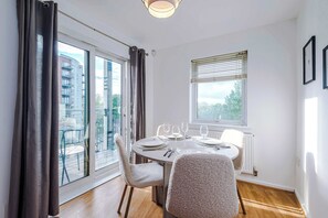 Un coin repas avec une table dressée pour quatre personnes, une fenêtre avec des stores et une vue sur un bâtiment à l’extérieur.