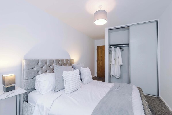 A bedroom with a tufted headboard, a bedside table with a lamp, a wardrobe, and a hanging robe.