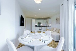 A modern dining area with a round table set for four, a kitchen in the background, and a sofa in the living area. - Spacious 2BR | Sleeps 6 | Parking | Walk to Etihad (Greater Manchester)