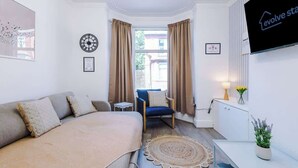 Ein modernes Hotelzimmer mit Bett, Sessel, Fernseher, Uhr und einem Fenster mit Vorhängen.