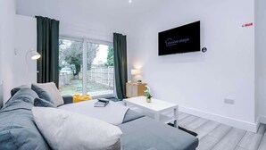 Ein modernes Wohnzimmer mit einer Couch, einem kleinen Tisch, einem an der Wand befestigten Fernseher und einer Glastür zum Außenbereich.