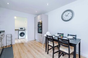 A modern kitchen with a microwave, oven, and washing machine, a dining table with chairs, and a wall clock.