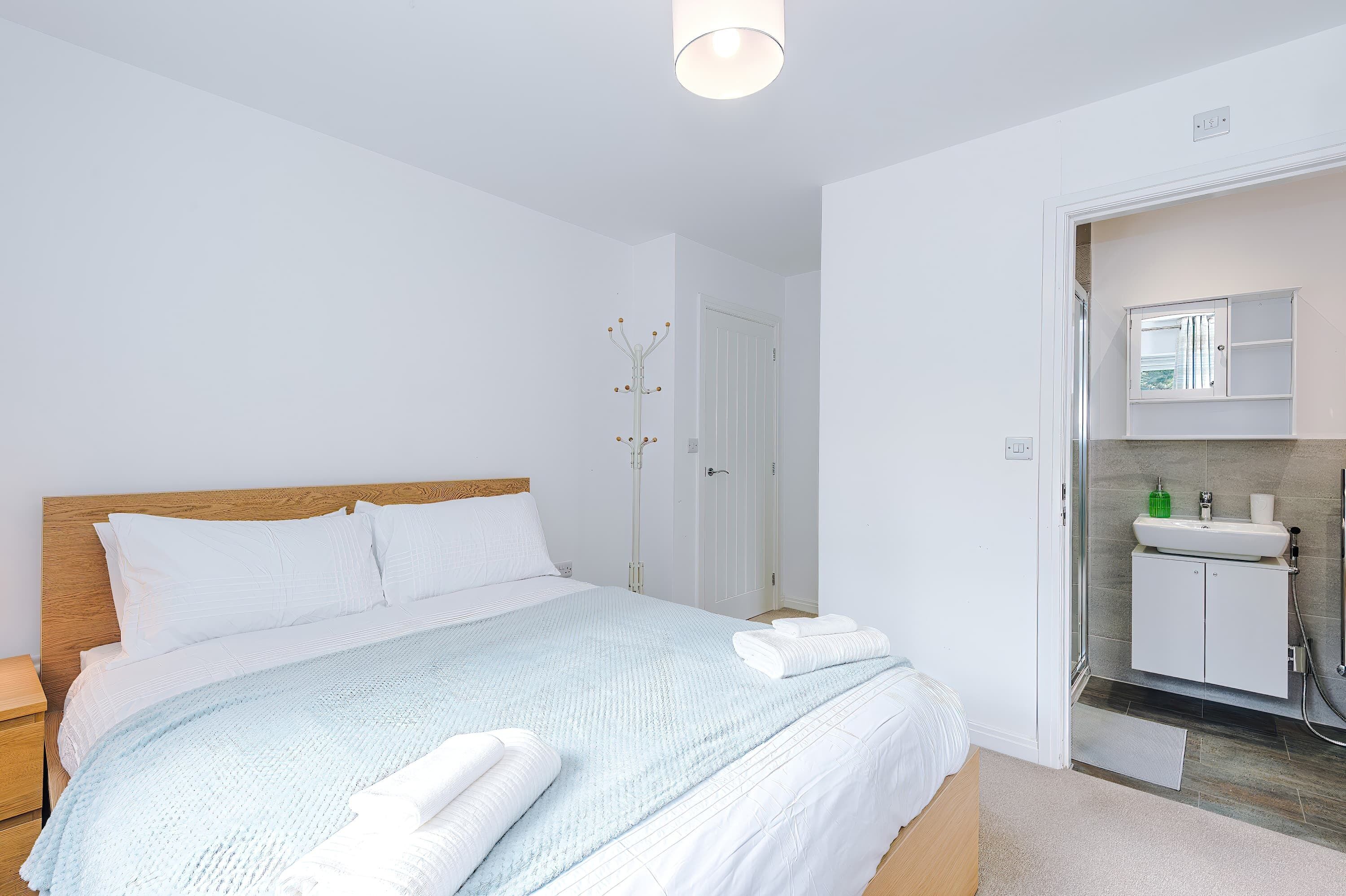 A bedroom with a bed, a nightstand, a bedside table, a bathroom with a sink, mirror, and shower.