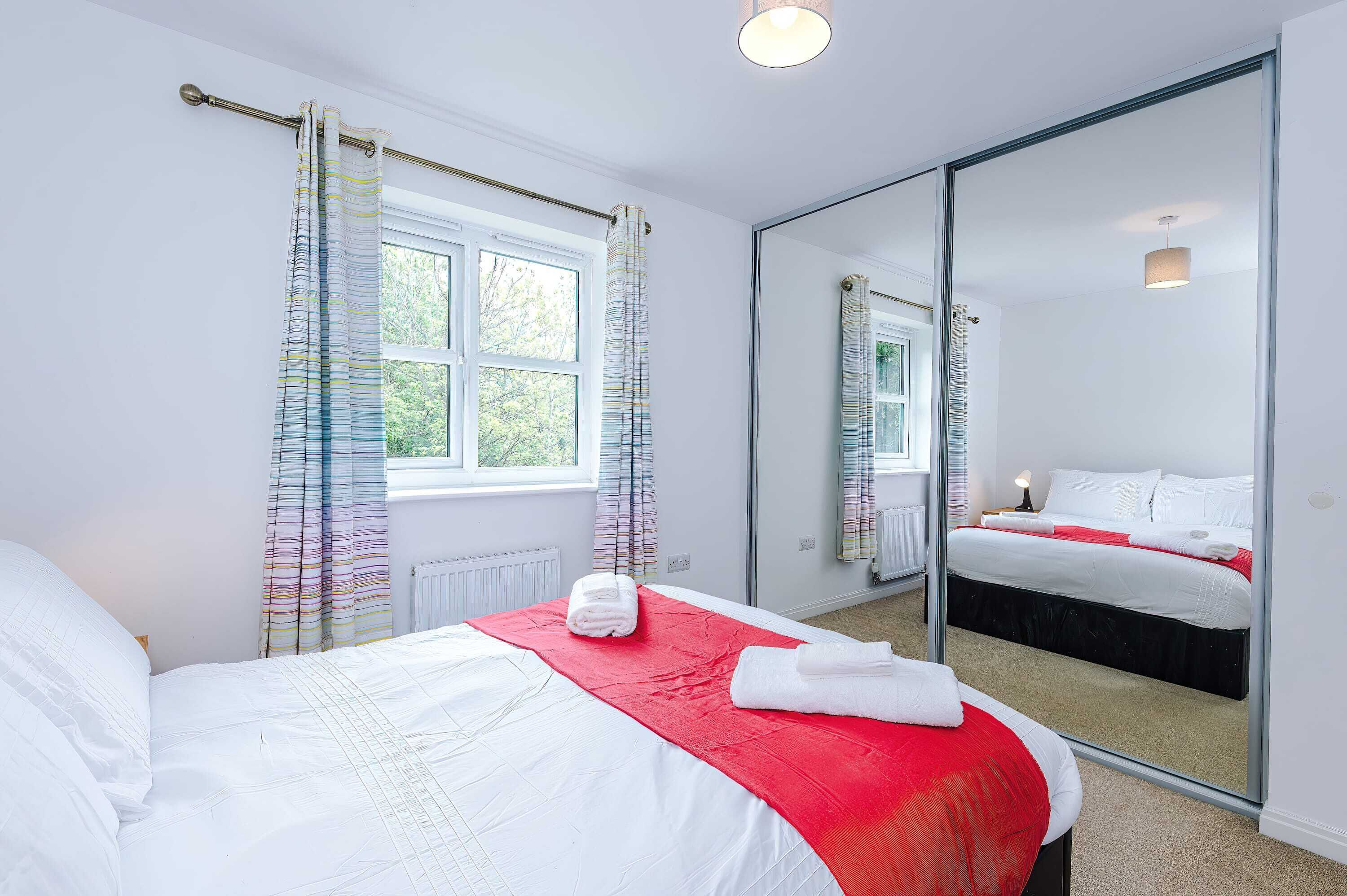 A hotel room with a large bed, a window with curtains, a mirror, and a bedside table with a lamp.